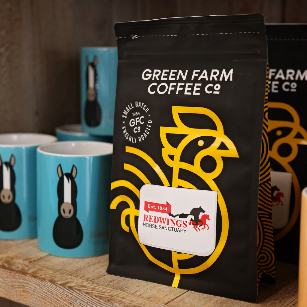 Redwings Horse Sanctuary Cafetiere Coffee. Roasted and ground by Green Farm Norfolk using their Dolce Vista blend. A black coffee bag featuring a yellow chicken and the Redwings Horse Sanctuary logo sitting on a wooden shelf at the Redwings Caldecott Visitor Centre shop.